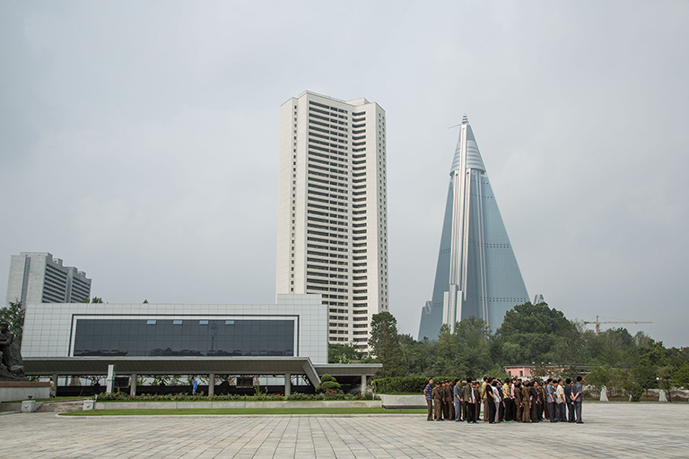 Typical occurrence: You arrive anyplace in NoKo; no one is there. Suddenly a swarm of people dressed in decades-old clothing arrives, organized in close formation, five or six people across, and as many deep, walking in lockstep. When I’d ask Older Handler why, she’d deny it was happening, even as we stood watching it. The infamous, pyramid-shaped Ryugyong Hotel can be seen in the background.